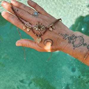 May include: A brown cord hand bracelet featuring a central gold-toned lotus flower charm with a small brown stone, accented by smaller gold-toned leaf charms and metallic beads, secured with thin adjustable cords.
