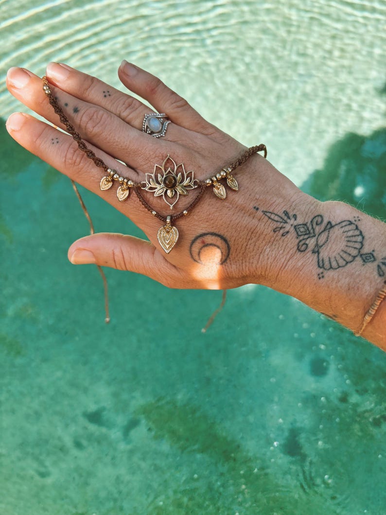 May include: A brown cord hand bracelet featuring a central gold-toned lotus flower charm with a small brown stone, accented by smaller gold-toned leaf charms and metallic beads, secured with thin adjustable cords.