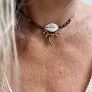 May include: A brown macrame choker necklace with a white seashell pendant. The necklace has gold-coloured accents and a gold leaf-shaped charm. The necklace is worn around the neck.
