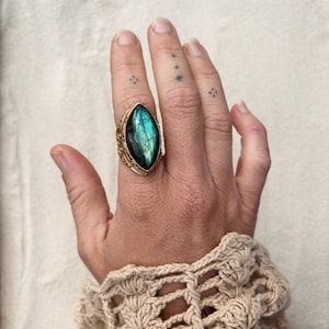 May include: A gold ring featuring a large, marquise-shaped labradorite gemstone with blue and green iridescence. The metal band has a textured, beaded edge surrounding the stone, creating a detailed frame for the polished, reflective surface.