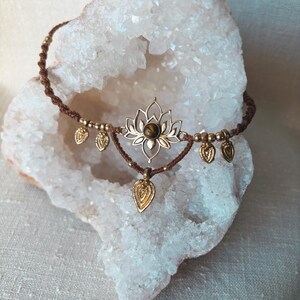 May include: A brown macrame choker necklace featuring a central gold-toned lotus flower charm with a round brown gemstone at its center. The necklace is adorned with small gold-toned beads and five dangling leaf-shaped charms along the cord.