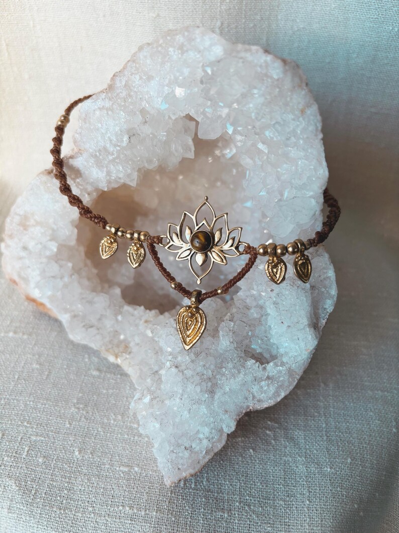 May include: A brown macrame choker necklace featuring a central gold-toned lotus flower charm with a round brown gemstone at its center. The necklace is adorned with small gold-toned beads and five dangling leaf-shaped charms along the cord.