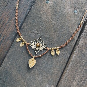 May include: A brown macrame cord necklace featuring a central gold-toned lotus flower pendant with a round brown gemstone at its center. The necklace is accented with small gold-toned beads and five dangling leaf-shaped charms in a matching finish.