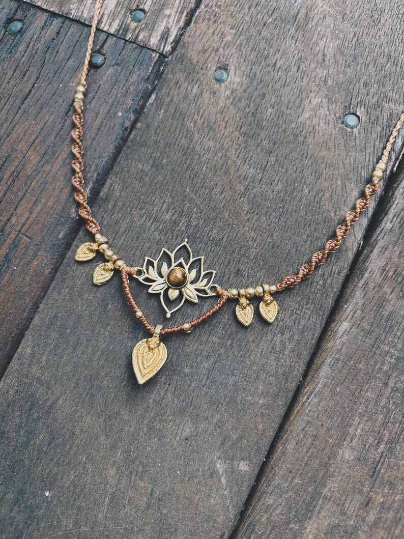 May include: A brown macrame cord necklace featuring a central gold-toned lotus flower pendant with a round brown gemstone at its center. The necklace is accented with small gold-toned beads and five dangling leaf-shaped charms in a matching finish.