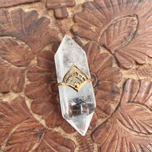 May include: A gold ring with a geometric design sits on a clear quartz crystal point. The crystal is resting on a wooden surface with a carved floral pattern.