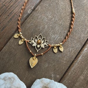 May include: A macrame necklace featuring a brown cord with a twisted design, accented by gold-toned metal beads and leaf-shaped charms. The centerpiece is a gold-toned lotus flower pendant with a central brown stone, and a dangling leaf charm.