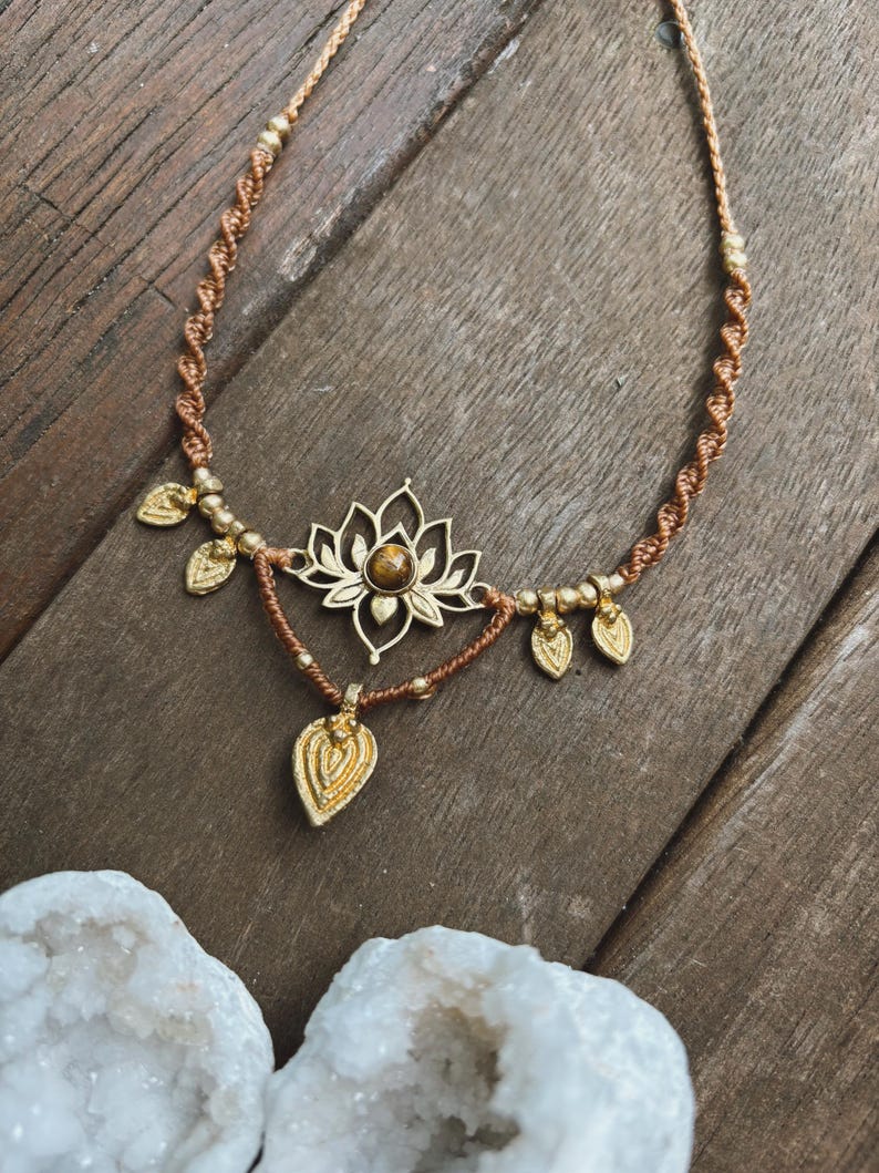 May include: A macrame necklace featuring a brown cord with a twisted design, accented by gold-toned metal beads and leaf-shaped charms. The centerpiece is a gold-toned lotus flower pendant with a central brown stone, and a dangling leaf charm.