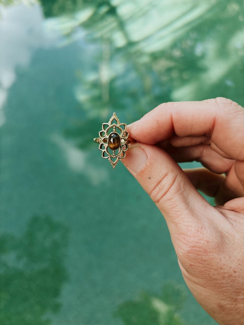 K&ouml;nnte beinhalten: Ein goldfarbener Ring mit einem komplizierten, symmetrischen, durchbrochenen Metallrahmen in Form einer Lotusbl&uuml;te. In der Mitte der dekorativen Metallarbeit befindet sich ein kleiner, glatter, ovaler brauner Edelstein mit einem dezenten Glanz.