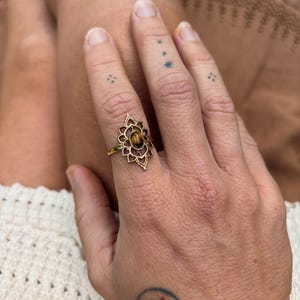 K&ouml;nnte beinhalten: Ein goldfarbener Ring mit einem ovalen Tigerauge-Edelstein, der in der Mitte eines komplizierten, symmetrischen Metallrahmens sitzt, wobei spitze, bl&uuml;tenblattartige Details von dem Stein nach au&szlig;en hin verlaufen.