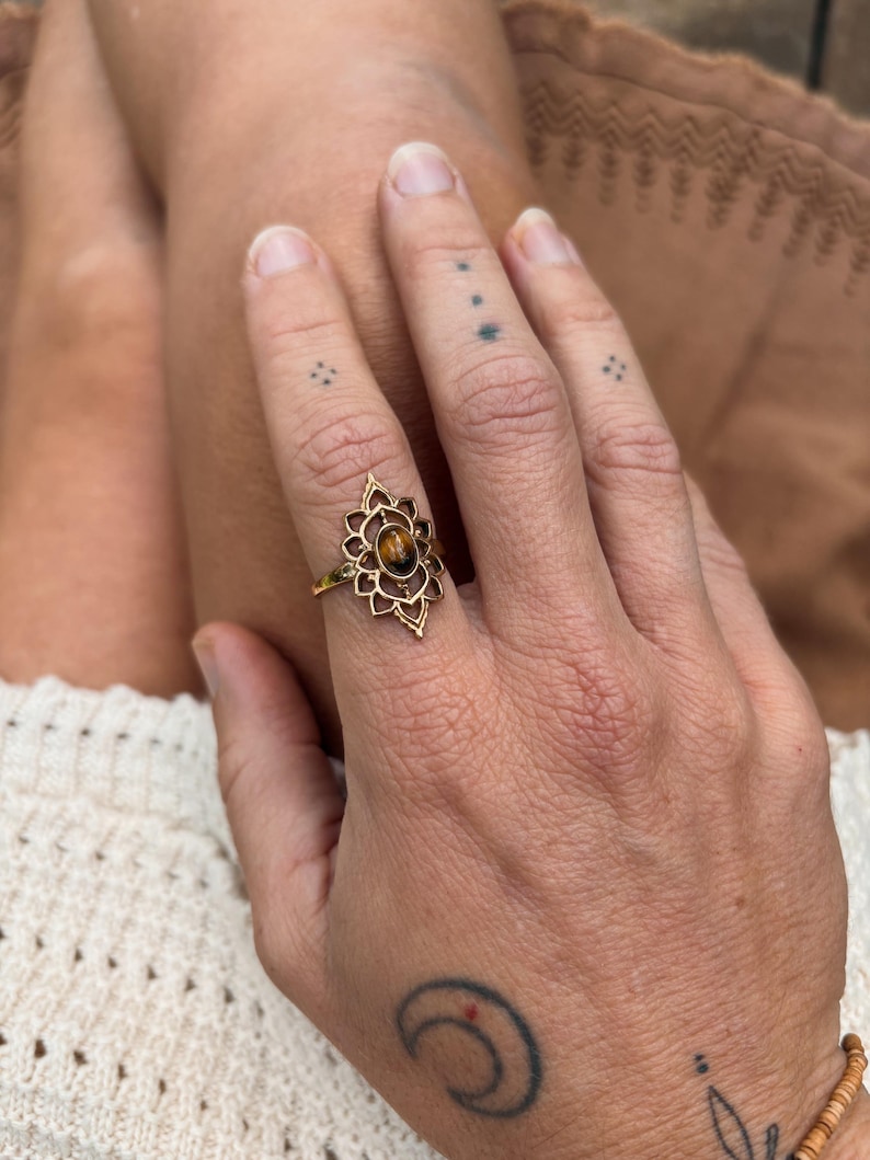 K&ouml;nnte beinhalten: Ein goldfarbener Ring mit einem ovalen Tigerauge-Edelstein, der in der Mitte eines komplizierten, symmetrischen Metallrahmens sitzt, wobei spitze, bl&uuml;tenblattartige Details von dem Stein nach au&szlig;en hin verlaufen.