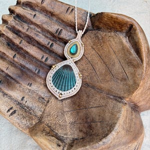 May include: A macrame pendant necklace featuring a large, dark teal, ribbed seashell and a smaller, teardrop-shaped, iridescent blue-green stone set in gold-toned metal. The stones are encased in light beige knotted cord with small gold-toned beads.