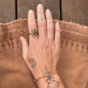 K&ouml;nnte beinhalten: Ein goldfarbener Ring mit einem ovalen, braunen Edelstein in einer kunstvollen, bl&uuml;tenblattartigen Metallfassung am Mittelfinger, kombiniert mit einem zarten Armband aus kleinen, zylindrischen beigen Perlen an einer d&uuml;nnen Metallschnur.