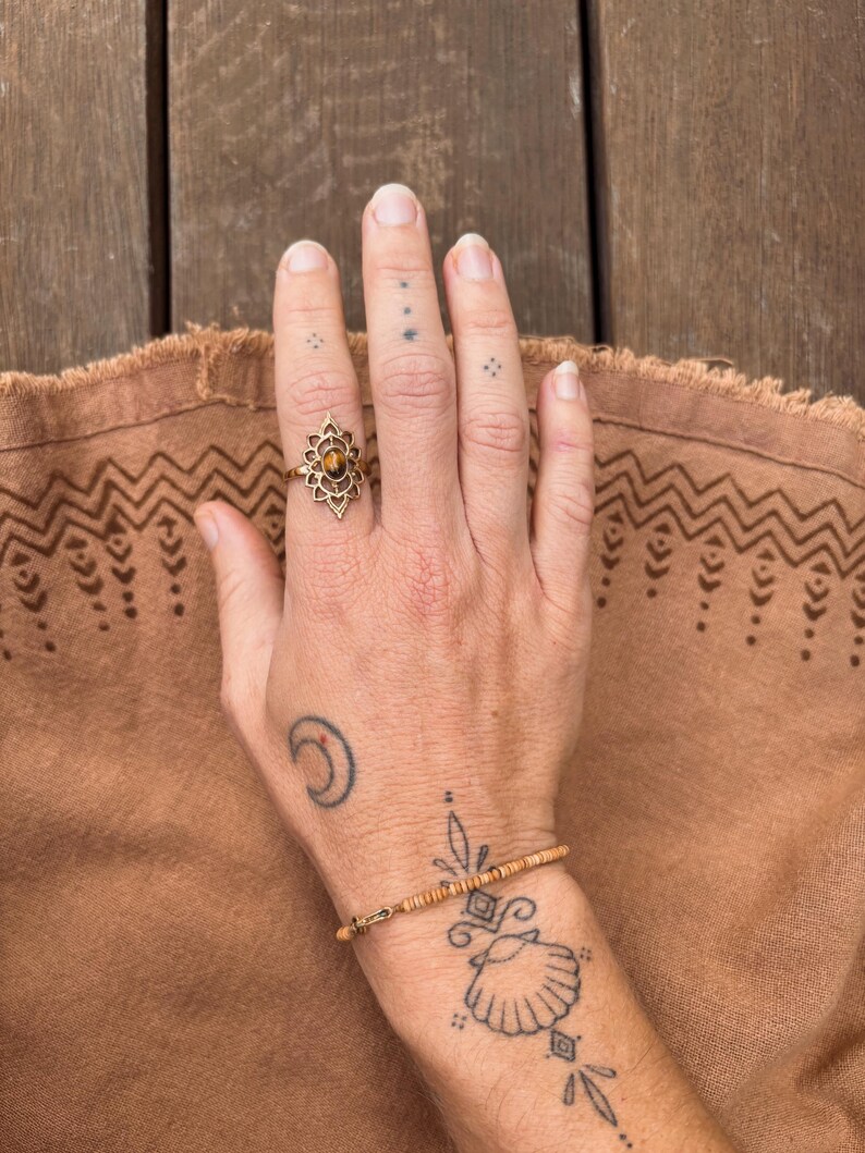 K&ouml;nnte beinhalten: Ein goldfarbener Ring mit einem ovalen, braunen Edelstein in einer kunstvollen, bl&uuml;tenblattartigen Metallfassung am Mittelfinger, kombiniert mit einem zarten Armband aus kleinen, zylindrischen beigen Perlen an einer d&uuml;nnen Metallschnur.