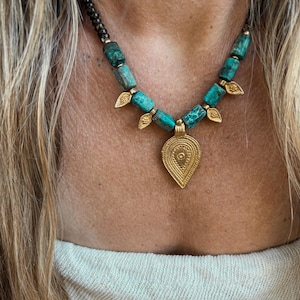 May include: A necklace featuring a central gold-toned teardrop pendant with intricate embossed patterns, flanked by turquoise-coloured cylindrical beads and small gold-toned leaf-shaped charms, strung on a dark beaded cord.