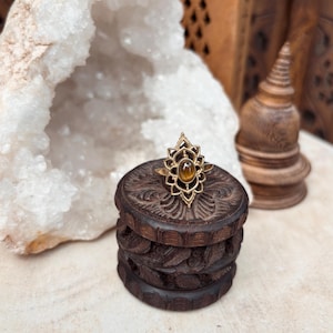 K&ouml;nnte beinhalten: Ein goldfarbener Ring mit einem ovalen, braunen Tigerauge-Edelstein, der in einen dekorativen, symmetrischen Metallrahmen eingefasst ist, welcher an eine stilisierte Lotusbl&uuml;te erinnert. Der Ring liegt auf einer kleinen, dunkelbraunen Holzschatulle.