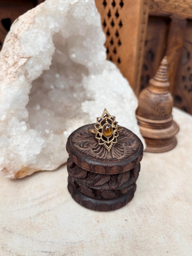K&ouml;nnte beinhalten: Ein goldfarbener Ring mit einem ovalen, braunen Tigerauge-Edelstein, der in einen dekorativen, symmetrischen Metallrahmen eingefasst ist, welcher an eine stilisierte Lotusbl&uuml;te erinnert. Der Ring liegt auf einer kleinen, dunkelbraunen Holzschatulle.