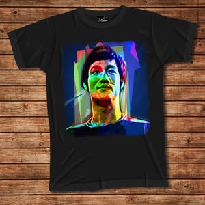 May include: A black short-sleeved t-shirt featuring a vibrant, multi-colored pop art style portrait of a person with dark hair. The graphic uses bold shades of blue, green, red, and yellow to create a stylized, angular, and abstract facial depiction.