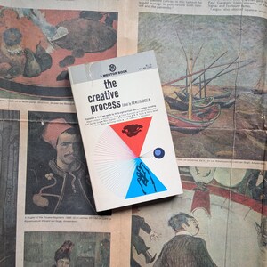 May include: A paperback book titled The Creative Process edited by Brewster Ghiselin, featuring a white cover with a grey top band, a red triangle, a blue triangle, a small blue circle, and black text. The book is positioned on a printed newspaper page.