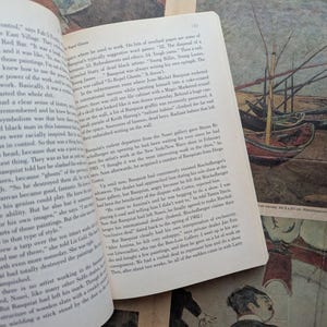 Puede incluir: Un libro abierto con texto en la p&aacute;gina izquierda y una pintura de barcos en la derecha. La pintura tiene una est&eacute;tica vintage, con un aspecto descolorido. El libro est&aacute; sobre una superficie estampada.
