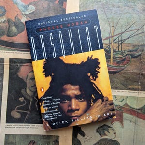 Puede incluir: Un libro de bolsillo titulado "Basquiat" de Phoebe Hoban. La portada presenta un retrato de Jean-Michel Basquiat con cabello negro sobre un fondo amarillo. El libro est&aacute; sobre un peri&oacute;dico antiguo con ilustraciones coloridas.