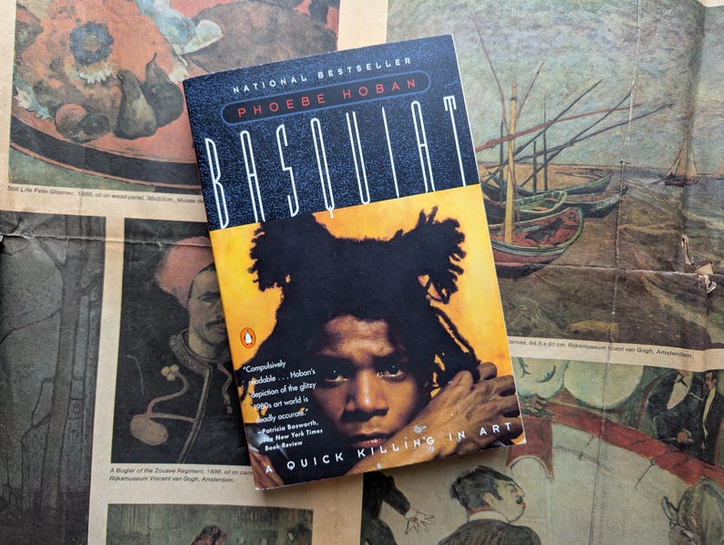 Puede incluir: Un libro de bolsillo titulado "Basquiat" de Phoebe Hoban. La portada presenta un retrato de Jean-Michel Basquiat con cabello negro sobre un fondo amarillo. El libro est&aacute; sobre un peri&oacute;dico antiguo con ilustraciones coloridas.