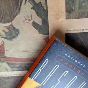 Puede incluir: Un libro de tapa dura con una cubierta azul oscuro y el t&iacute;tulo "RACE" en blanco y el nombre del autor "PHOEBE HOBAN" en rojo. El libro es parcialmente visible, descansando sobre una impresi&oacute;n vintage con obras de arte.