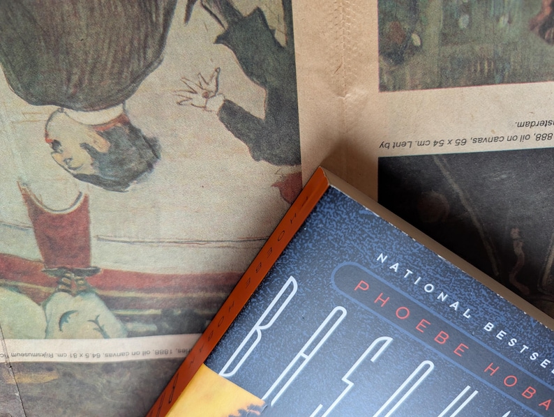 Puede incluir: Un libro de tapa dura con una cubierta azul oscuro y el t&iacute;tulo "RACE" en blanco y el nombre del autor "PHOEBE HOBAN" en rojo. El libro es parcialmente visible, descansando sobre una impresi&oacute;n vintage con obras de arte.