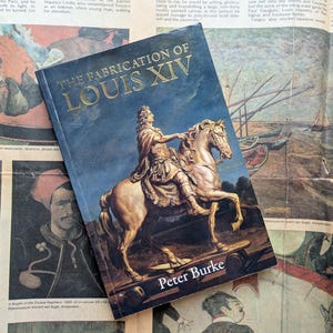 May include: A book titled "The Fabrication of Louis XIV" by Peter Burke. The cover features a gold statue of a person on a horse against a dark blue background. The book is resting on a newspaper with various images.