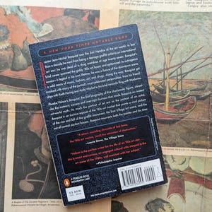 Puede incluir: Una biograf&iacute;a de Jean-Michel Basquiat de Penguin Books, un libro notable del New York Times. La portada del libro es negra con texto blanco y un logotipo de Penguin rojo. El libro descansa sobre un peri&oacute;dico antiguo con obras de arte coloridas.