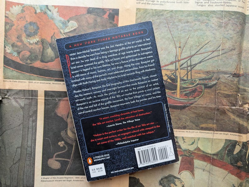 Puede incluir: Una biograf&iacute;a de Jean-Michel Basquiat de Penguin Books, un libro notable del New York Times. La portada del libro es negra con texto blanco y un logotipo de Penguin rojo. El libro descansa sobre un peri&oacute;dico antiguo con obras de arte coloridas.
