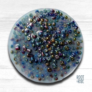 May include: A circular, light blue resin piece filled with a variety of iridescent beads. The beads are in shades of blue, purple, gold, and green. The piece is on a white wooden surface. The Root & Rise logo is visible.