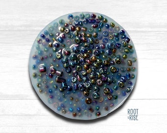 May include: A circular, light blue resin piece filled with a variety of iridescent beads. The beads are in shades of blue, purple, gold, and green. The piece is on a white wooden surface. The Root & Rise logo is visible.