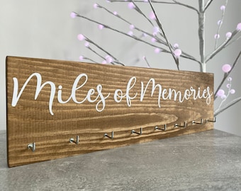 Könnte beinhalten: Ein Holzschild mit dem Text "Miles of Memories" in weißen Buchstaben. Das Schild hat neun Haken zum Aufhängen von Gegenständen.