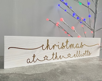 Puede incluir: Un letrero de madera blanco con letras doradas que dice "Christmas at the Elliots". El letrero tiene una textura de grano de madera rústico y está decorado con estrellas doradas.