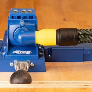 May include: A blue Kreg pocket hole jig with a yellow and black hose attached. The jig is mounted on a light brown wooden surface. The jig has a black handle and the Kreg logo is visible.