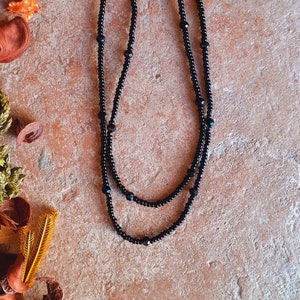 Puede incluir: Un collar negro de cuentas en capas con cuentas pequeñas y grandes. El collar tiene un diseño delicado con algunas cuentas de color turquesa intercaladas. El fondo es una superficie texturizada de color marrón rojizo con hojas y plantas secas.