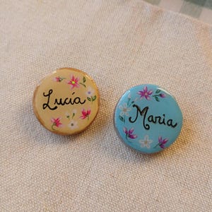 Puede incluir: Dos broches circulares de madera con diseños florales pintados a mano. El broche izquierdo es de color amarillo claro con el nombre Lucia escrito en letra negra. El derecho es azul claro con el nombre Maria en negro, rodeado de flores rosas y blancas.