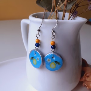 May include: A pair of handmade earrings featuring a light blue circular design with a painted lemon and white flower. The earrings have silver-tone hooks and are accented with orange and blue beads.