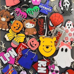 May include: A collection of assorted silicone keychains featuring various designs including a pink scrub top, a ghost character, a yellow bear face, a pumpkin, a Spider-Man mask, a Coca-Cola logo, a Highland cow, and text labels like LPN, RN, and CNA.