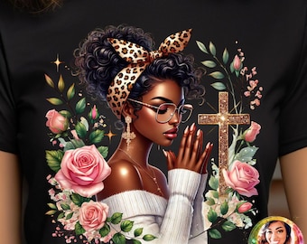 May include: A black t-shirt with a graphic of a woman with brown skin praying with a gold cross and pink flowers. The woman is wearing a leopard print headband and glasses. The text "KB Designs LLC" is in the bottom right corner.