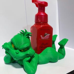 May include: A bright green Grinch-shaped plastic holder for a red Bath & Body Works foaming soap dispenser. The soap bottle features a graphic of a red sleigh with gifts and the text Bright Christmas Morning in a cursive font.