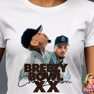 May include: White t-shirt featuring a graphic of two men wearing baseball caps. The graphic includes the text "BREEZY BOWL Chris Brown XX" in brown and black. The shirt also has a small logo in the bottom right corner.