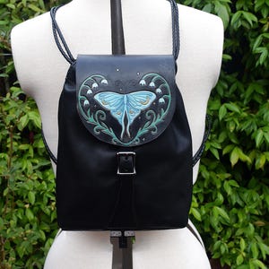 May include: A black backpack featuring a flap with a hand-painted light blue moth and white lily of the valley floral design. The bag includes a silver-toned metal buckle closure and thin black braided straps, displayed on a white mannequin.