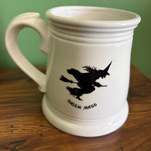 May include: A cream-colored ceramic mug featuring a black silhouette graphic of a witch flying on a broomstick. Below the graphic, the text reads SALEM MASS in black capital letters. The mug has a wide handle and a raised rim at the top and base.