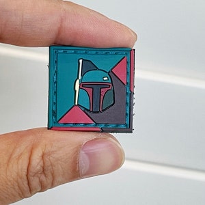 May include: A small, square patch with a black, green, and red geometric design. The center of the patch features a stylized image of a Star Wars character's helmet, Boba Fett.