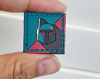 Può includere: Un piccolo patch quadrato con un design geometrico in nero, verde e rosso. Al centro del patch, un'immagine stilizzata del casco di un personaggio di Star Wars, Boba Fett.