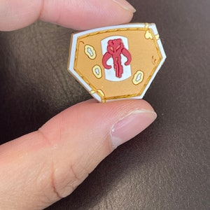 Könnte beinhalten: Eine kleine, sechseckige, goldfarbene Anstecknadel mit einem roten und weißen Mandalorianer-Symbol in der Mitte. Die Anstecknadel hat einen weißen Rand und eine goldfarbene Umrandung.