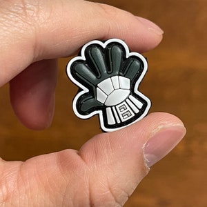 May include: Black and white rubber pin of a hand wearing a goalie glove. The glove has a white design with the letters "5G" on the wrist.