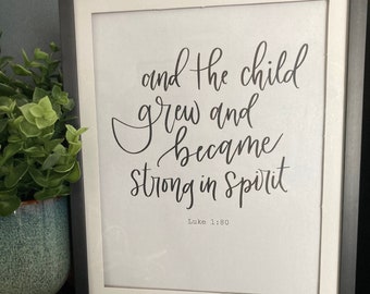 May include: A black framed print with a white background and black calligraphy text that reads "and the child grew and became strong in spirit" with the scripture reference "Luke 1:80" below the text.