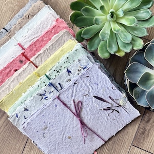 Puede incluir: Una pila de papel hecho a mano en varios colores, incluyendo rosa, amarillo y lavanda, atada con una cuerda rosa. El papel tiene un aspecto texturizado y orgánico con inclusiones visibles de flores secas y materia vegetal. Suculentas en el fondo.